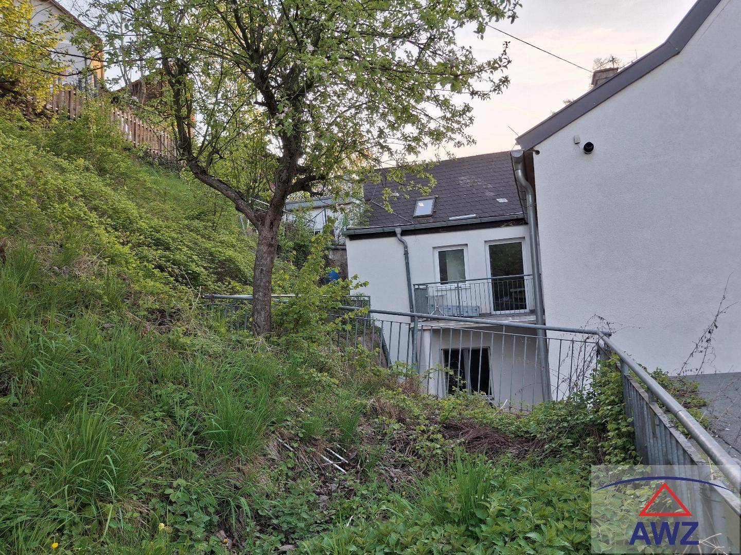 Ansicht des Hauses von der Seite, umgeben von üppigem Grün und einem Hang mit Bäumen.