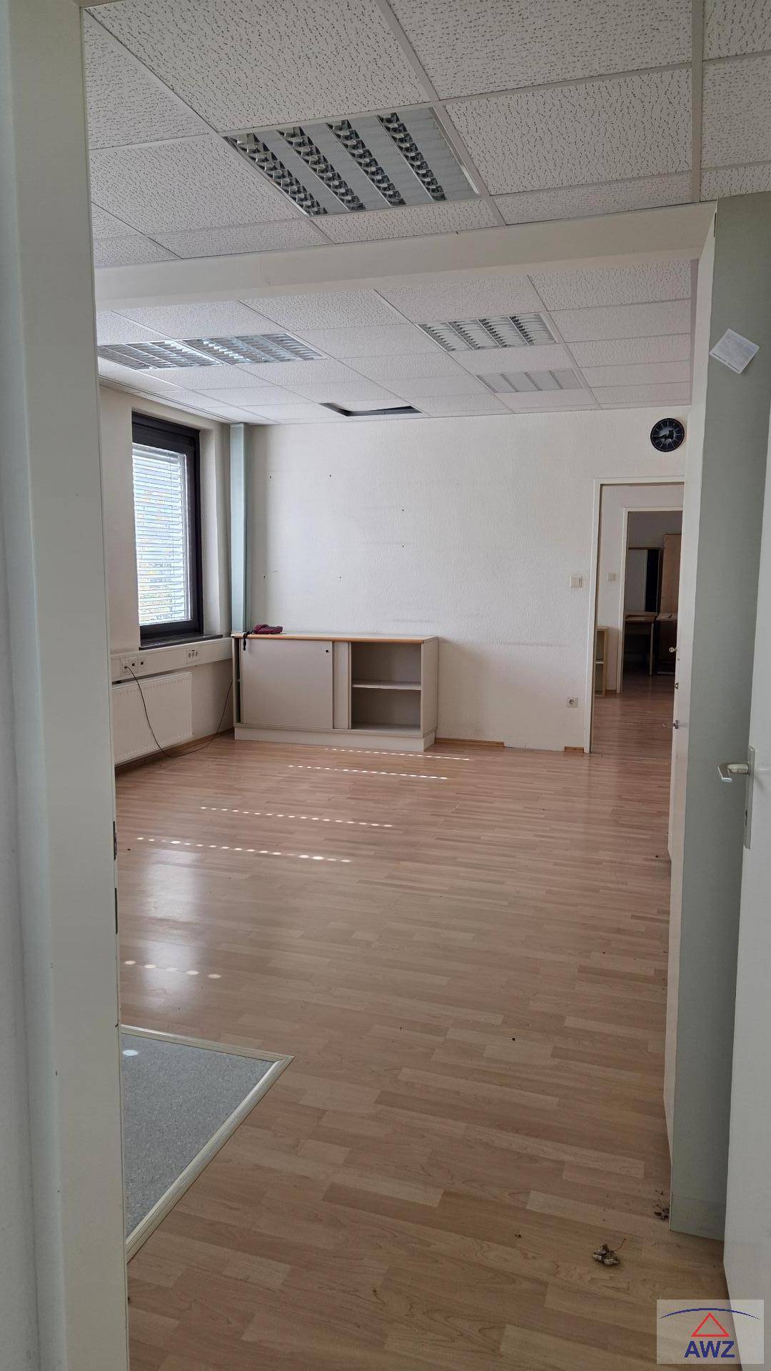 Heller Büroraum mit Laminatboden, abgehängter Decke und großem Fenster.