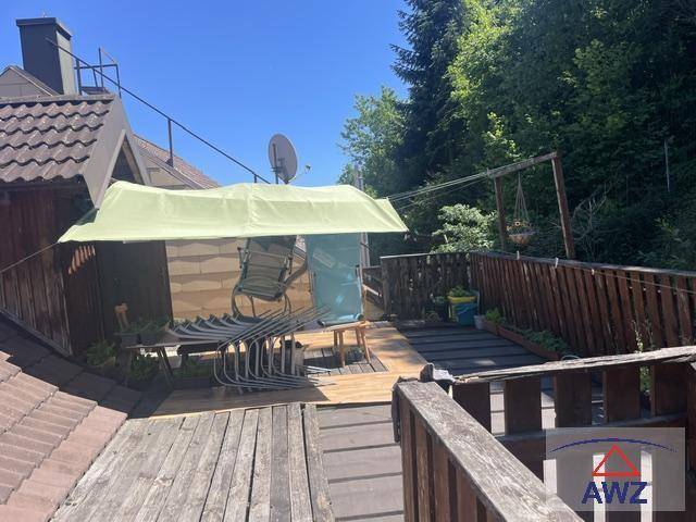 Sonniger Terrassenbereich mit Holzdeck, Sitzgelegenheiten und grüner Überdachung, umgeben von Bäumen.