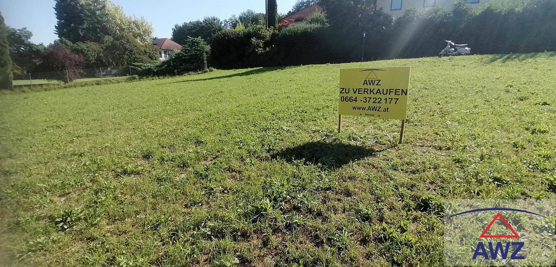 Sonnige Grünfläche mit einem 'Zu verkaufen'-Schild, umgeben von Bäumen und einem Haus im Hintergrund.