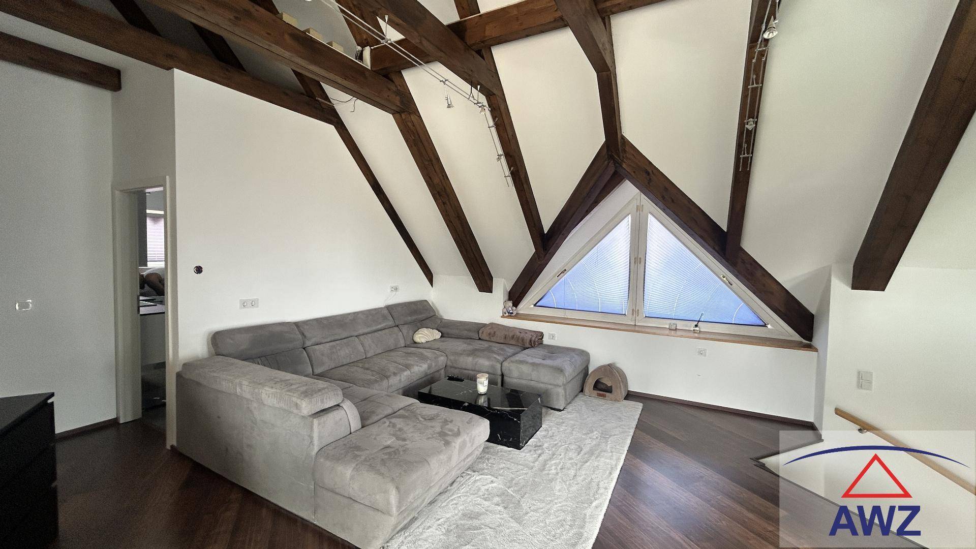 Gemütliches Wohnzimmer im Dachgeschoss mit freiliegenden Holzbalken, großem Sofa und dreieckigem Fenster.