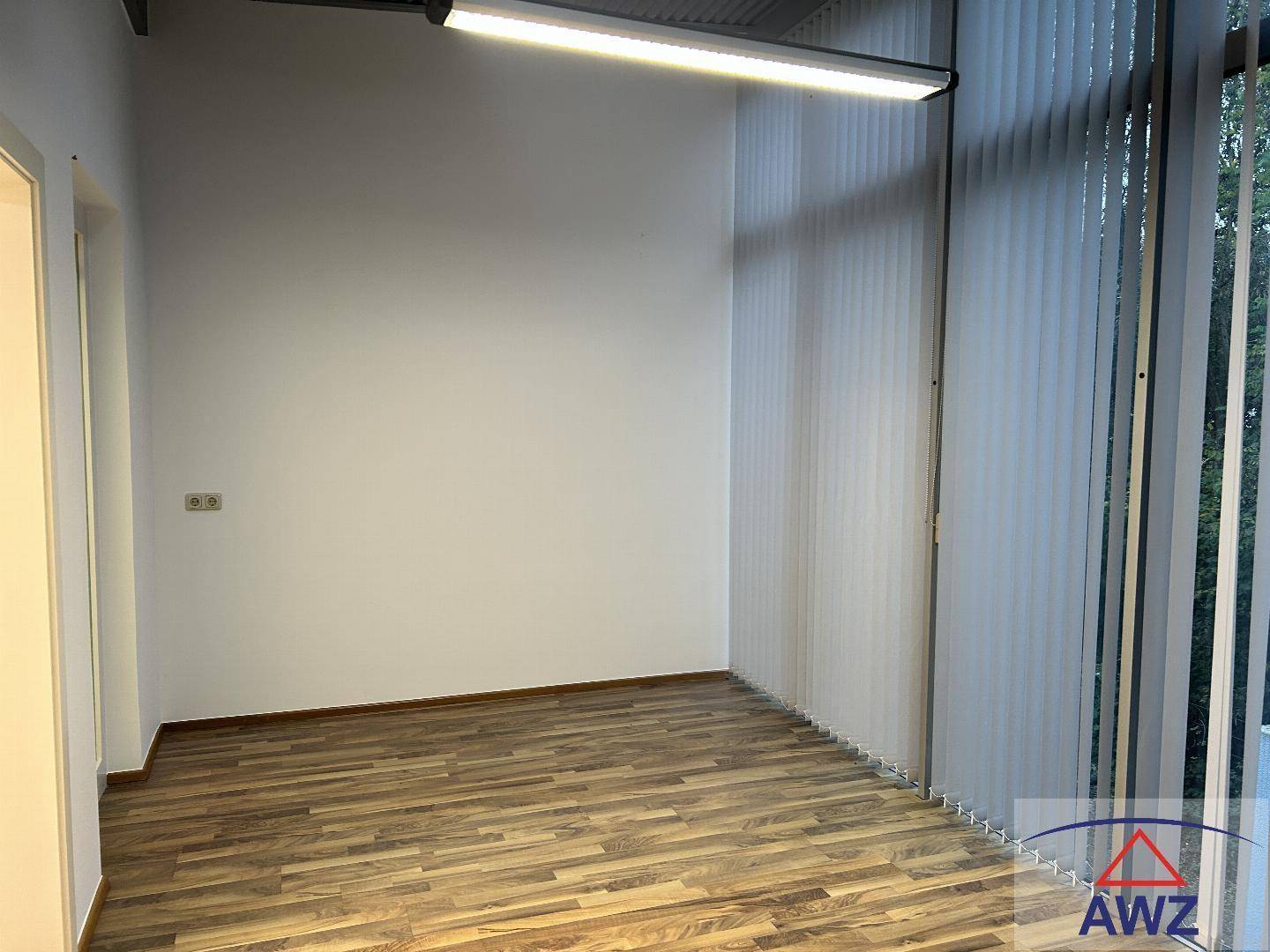 Heller leerer Raum mit Laminatboden und großen Fenstern mit vertikalen Jalousien.