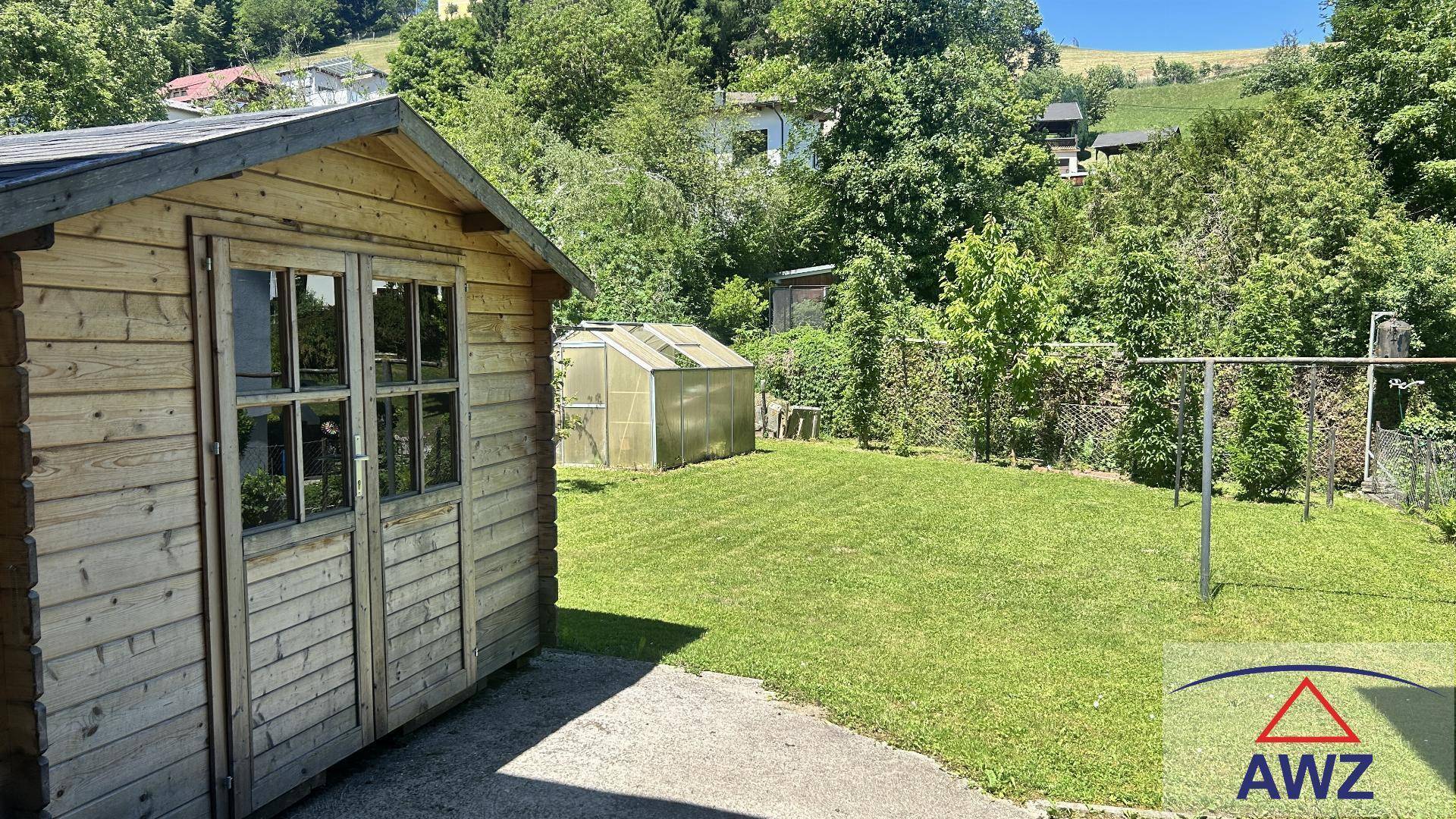 Gepflegter Gartenbereich mit einem Holzschuppen, Rasenfläche und einem kleinen Gewächshaus im Hintergrund.