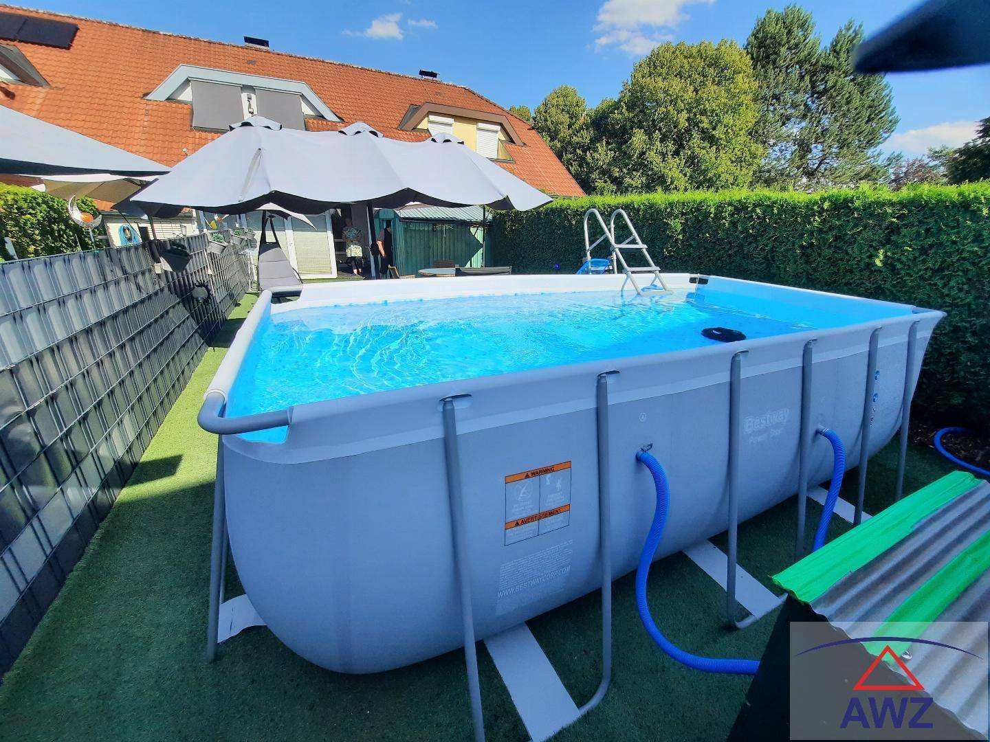 Großer Aufstellpool im Garten mit Leiter und blauer Wasseroberfläche unter blauem Himmel.