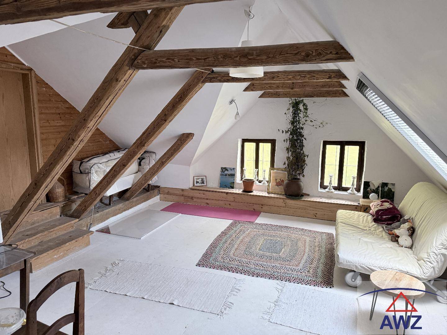 Helles Dachzimmer mit sichtbaren Holzbalken und schrägen Wänden, eingerichtet als gemütlicher Wohnbereich.