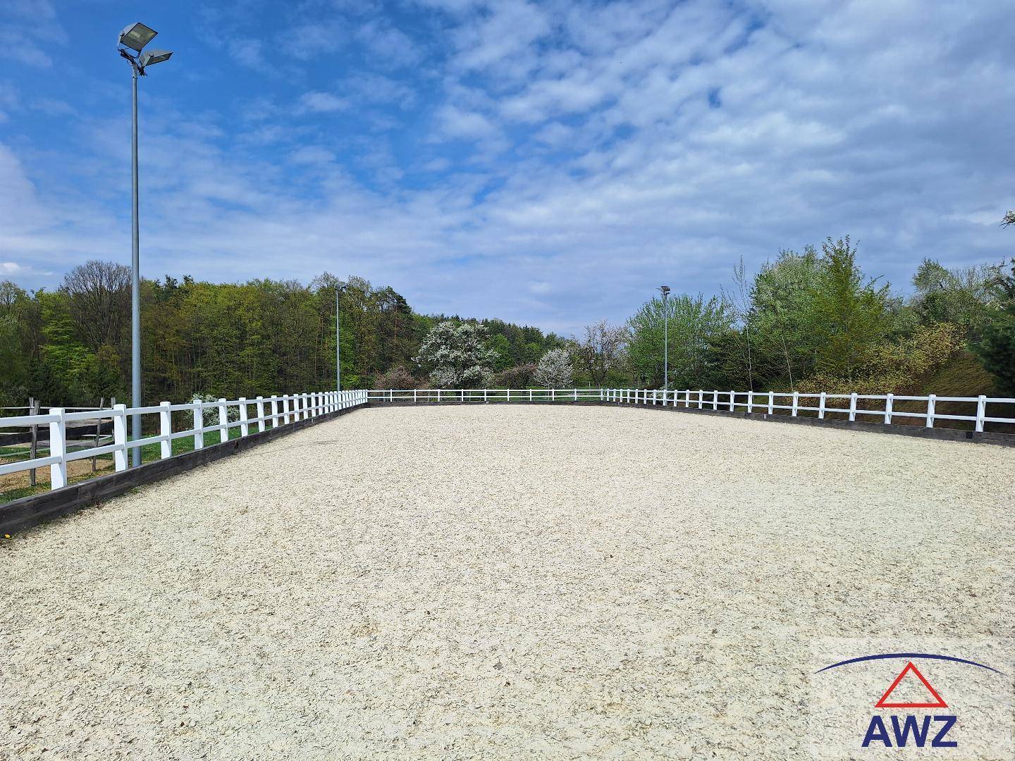 Großer Reitplatz im Freien mit weißem Zaun und grüner Umgebung unter blauem Himmel.