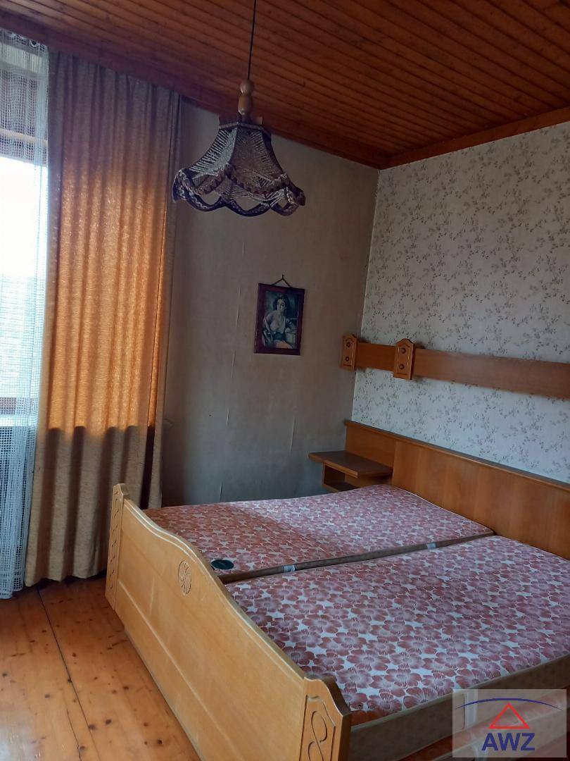 Schlafzimmer mit Holzdoppelbett, Holzdecke und gemusterter Tapete, das durch ein Fenster mit Vorhängen erhellt wird.