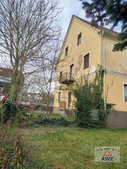 Gelbes Gebäude mit Satteldach, kleinem Balkon und üppiger Vegetation, umgeben von einem großen, kahlen Baum.