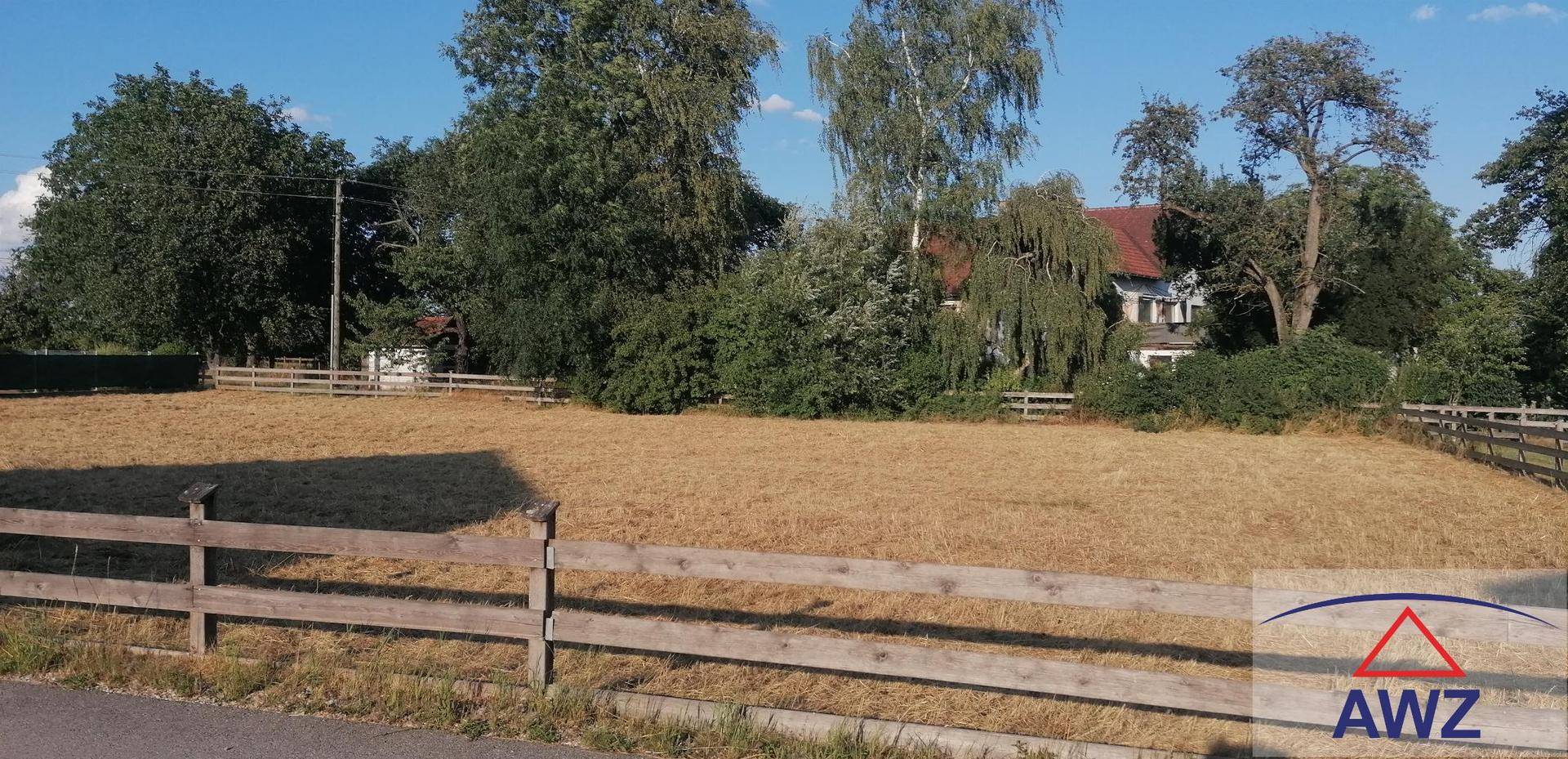Sonniges Baugrundstück mit Holzzaun, umgeben von Bäumen und einem Haus in der Ferne.