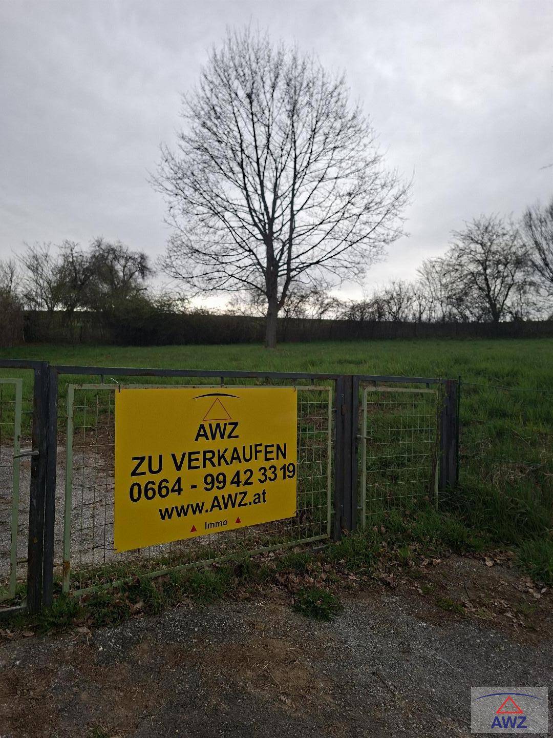 Eingang zu einem Grundstück mit einem gelben Verkaufsschild an einem Metalltor und weitem Blick ins Grüne.