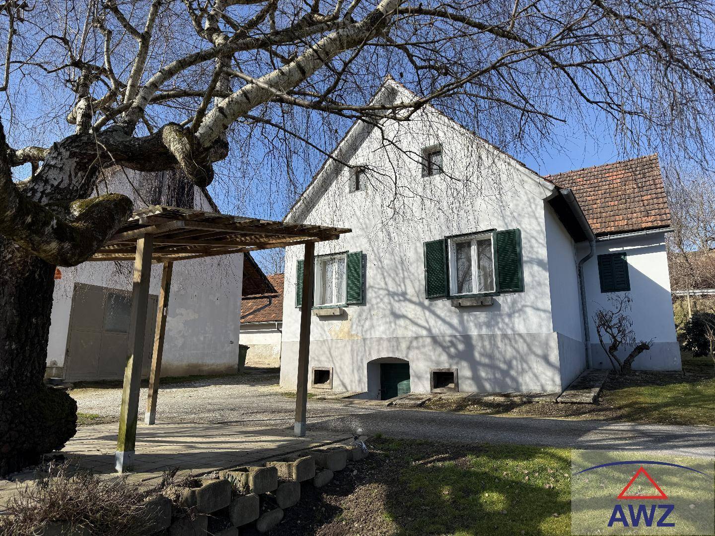 Landhausansicht mit einem einfachen Carport und einem großen, alten Baum im Vordergrund.