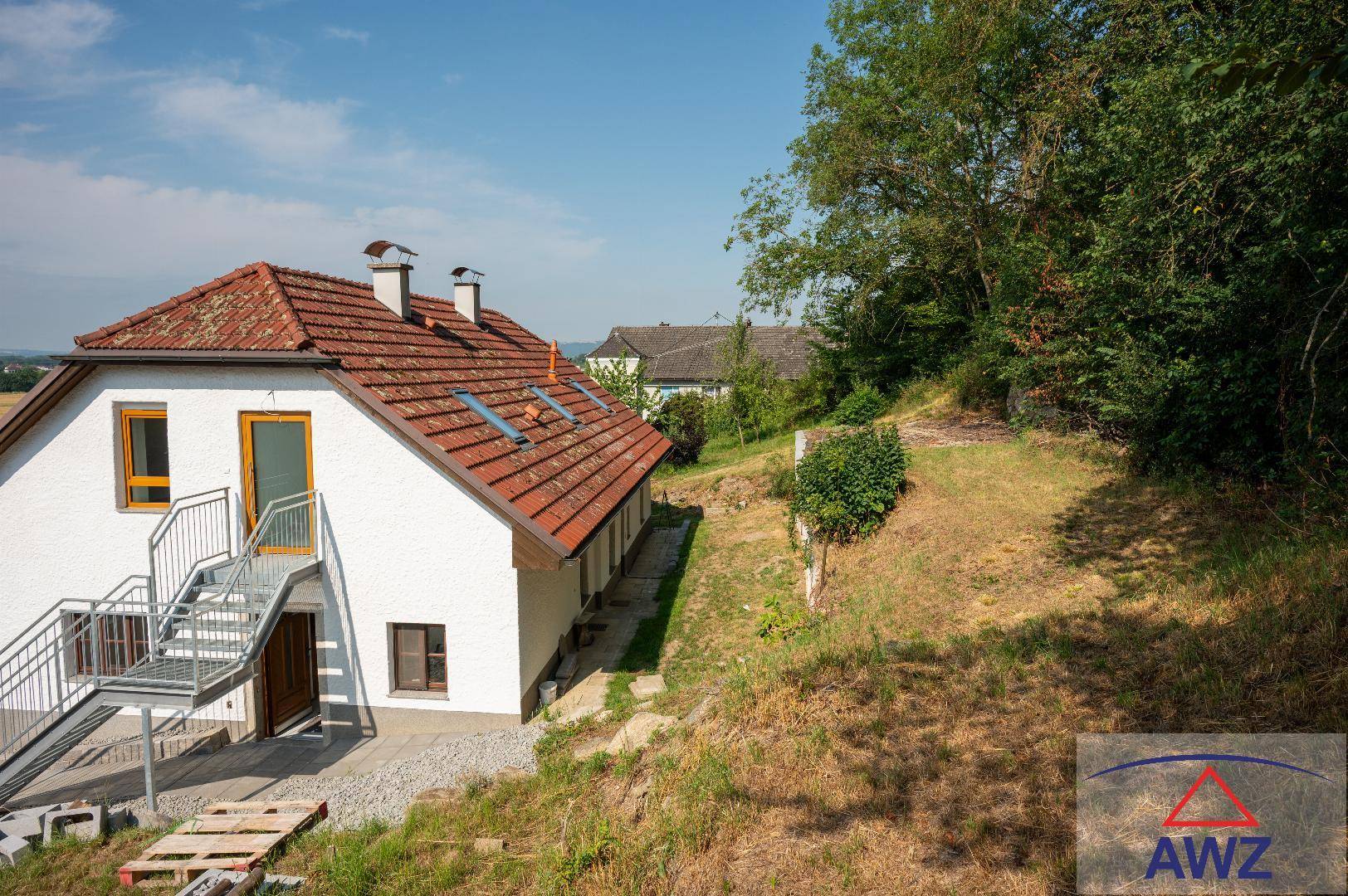 Hausfassade mit Außentreppe und angrenzendem, leicht abfallendem Gartenbereich unter blauem Himmel.