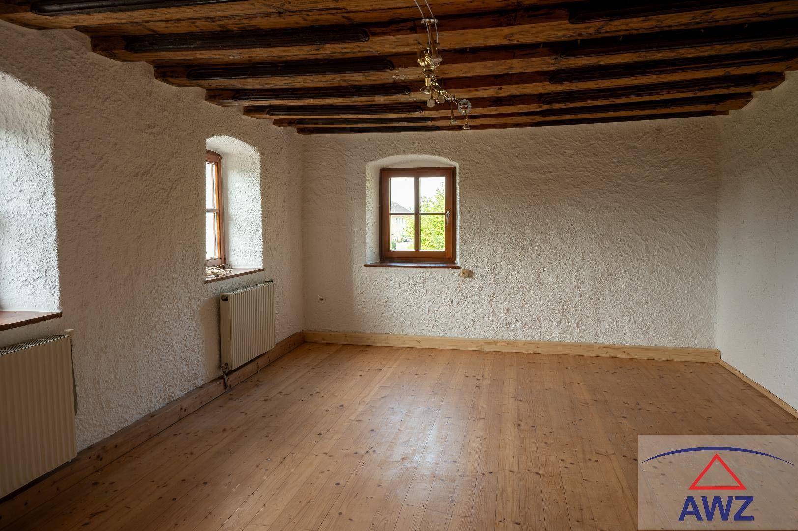 Großer Raum mit rustikaler Holzbalkendecke, Holzboden und zwei Fenstern.