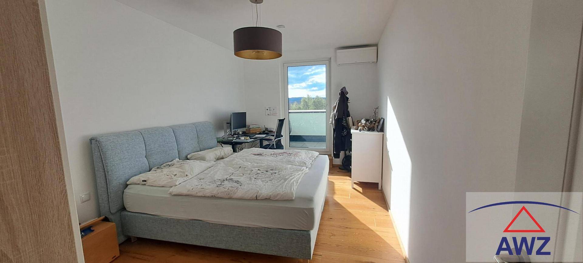 Schlafzimmer mit Doppelbett, Schreibtisch und Zugang zu einem Balkon, ausgestattet mit Holzfußboden.