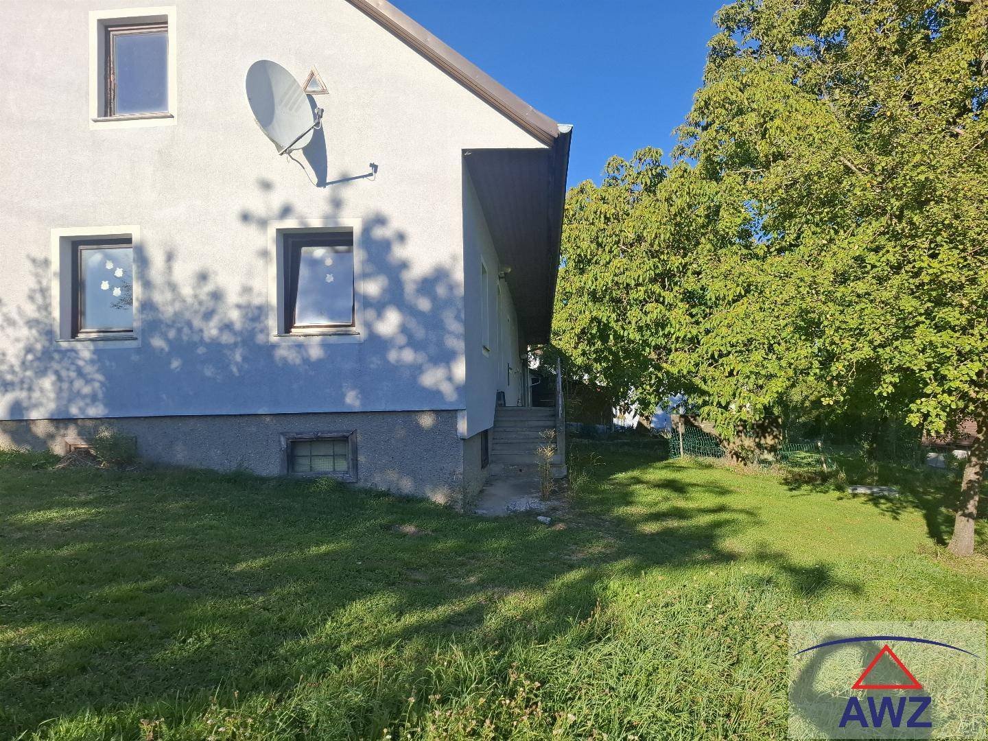 Seitenansicht des Hauses mit Satellitenschüssel und großem Baum im sonnigen Garten.