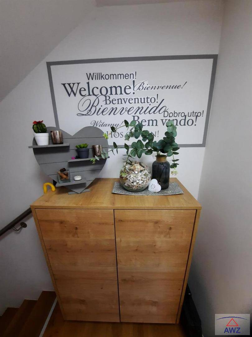 Heller Eingangsbereich mit Holzschrank, dekorativen Pflanzen und einer mehrsprachigen Willkommenstafel.