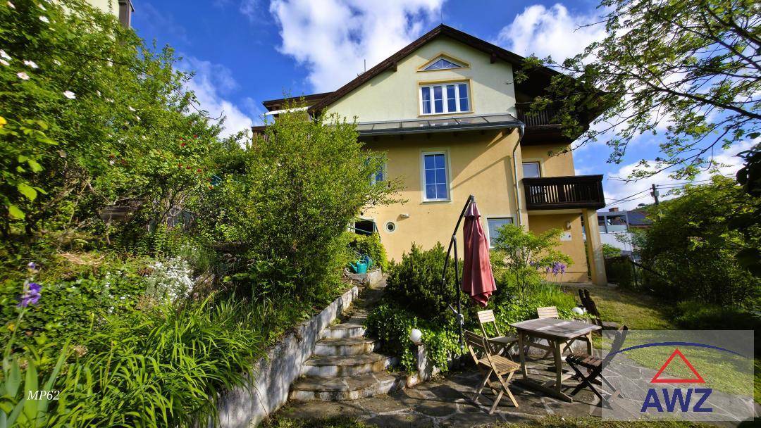 Hausfassade umgeben von einem grünen Garten mit Sitzbereich und Steintreppen.