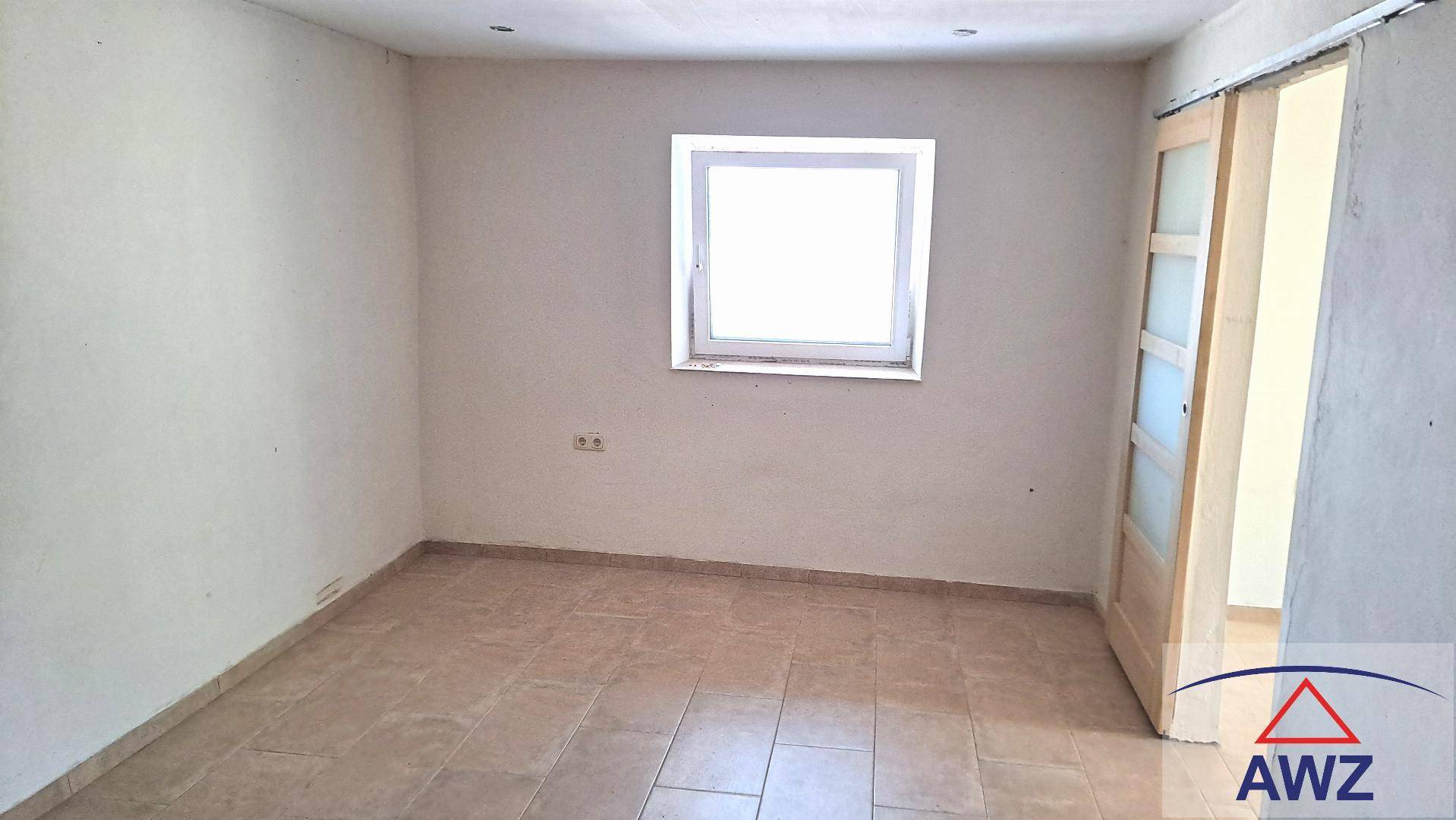 Leerer Raum mit Fliesenboden, weißen Wänden und einem Fenster, mit einer offenen Schiebetür aus Holz.