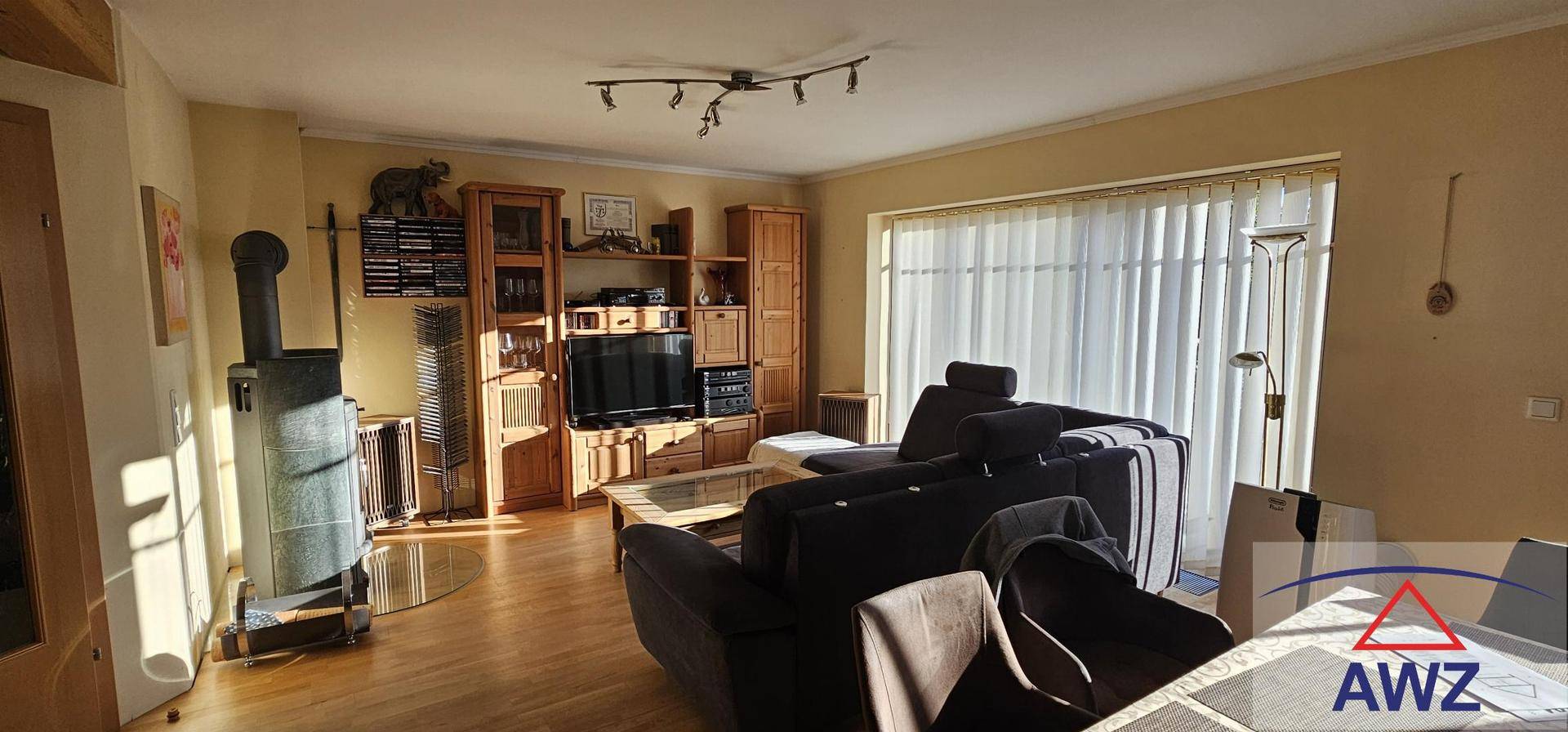 Gemütliches Wohnzimmer mit Holzofen, Parkettboden und großem Fenster mit Jalousien.