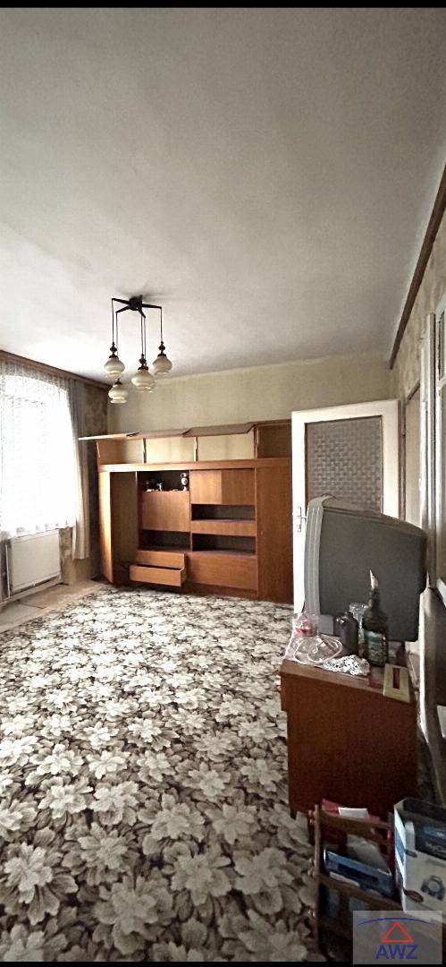 Wohnzimmer mit einem floral gemusterten Teppichboden, Holzmöbeln und einem Fenster mit Heizkörper.