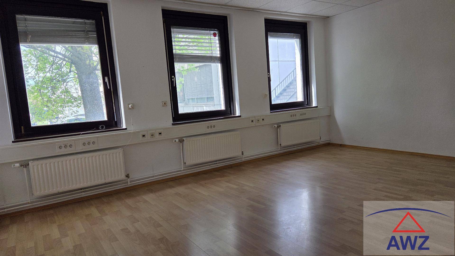 Leerer Büroraum mit Holzboden, großen Fenstern und Heizkörpern unter den Fenstern, bietet viel Tageslicht.