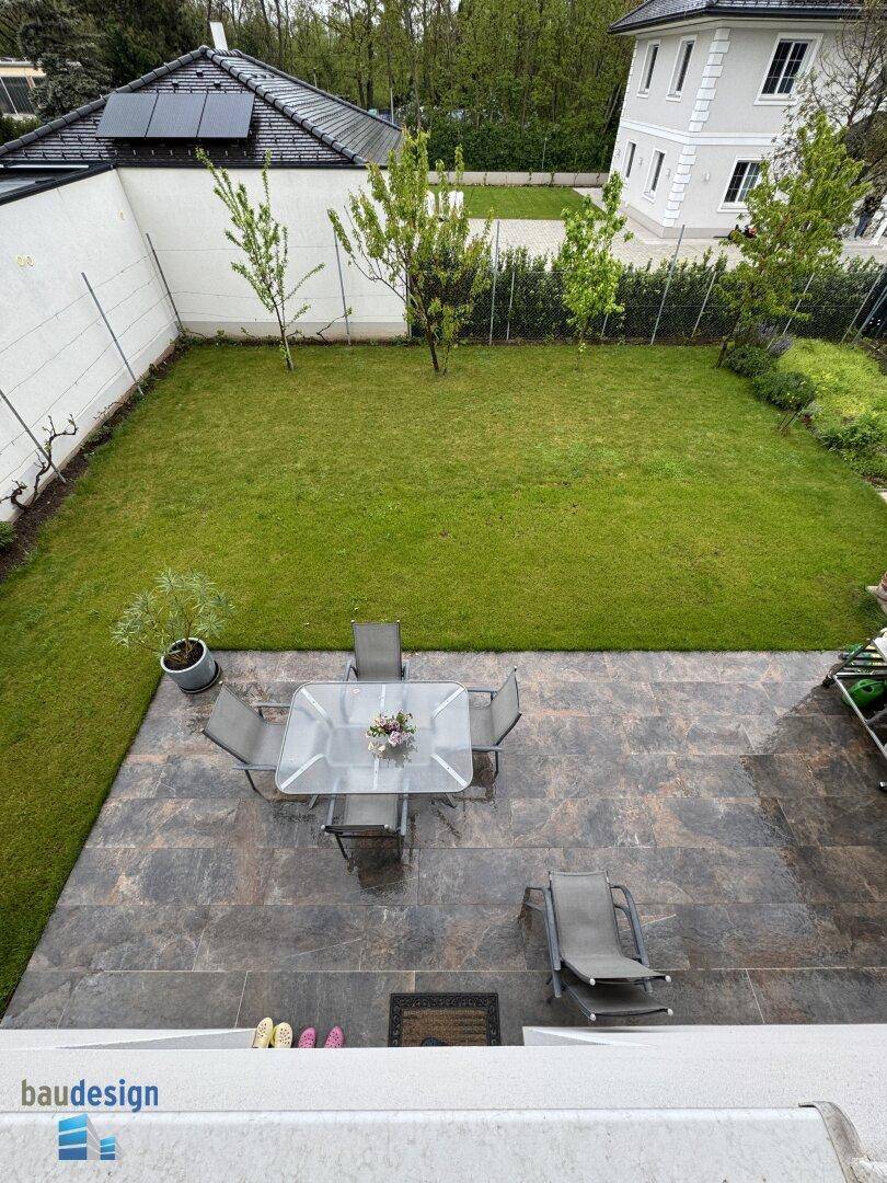 Blick von oben auf eine geflieste Terrasse mit Gartenmöbeln und gepflegtem Rasenbereich.