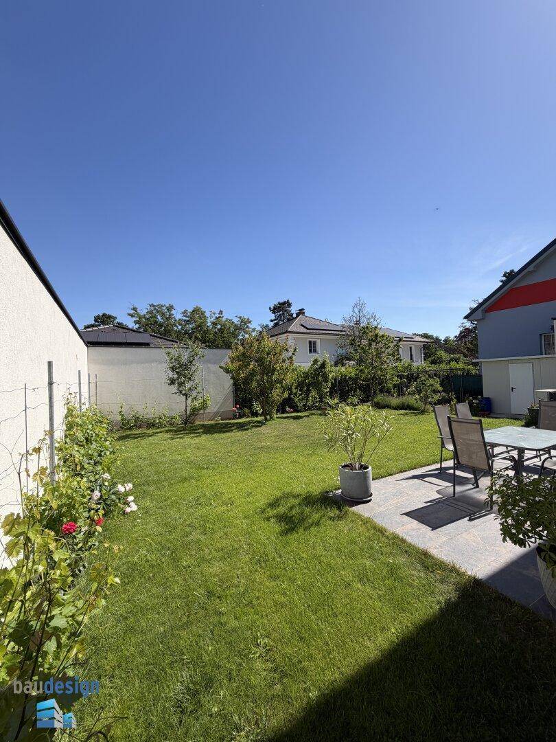 Sonniger Gartenbereich mit Rasenfläche, Terrasse und umliegender grüner Bepflanzung.