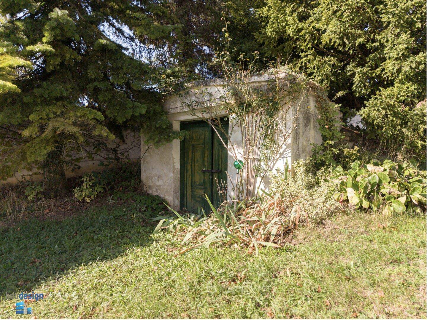 Kleines, altes Gebäude mit grüner Holztür, umgeben von Bäumen und Sträuchern im Garten.