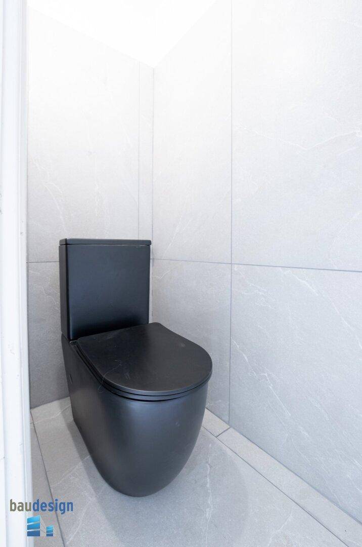 Modernes WC mit schwarzer Toilette und hellgrauen Fliesen an Wänden und Boden.