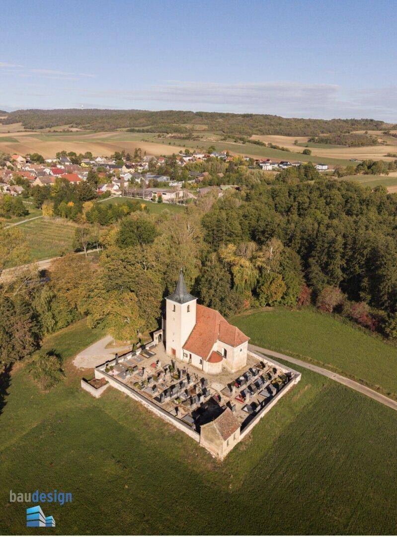 Luftaufnahme einer Kirche mit Friedhof, umgeben von Feldern und einem Dorf in der Ferne.