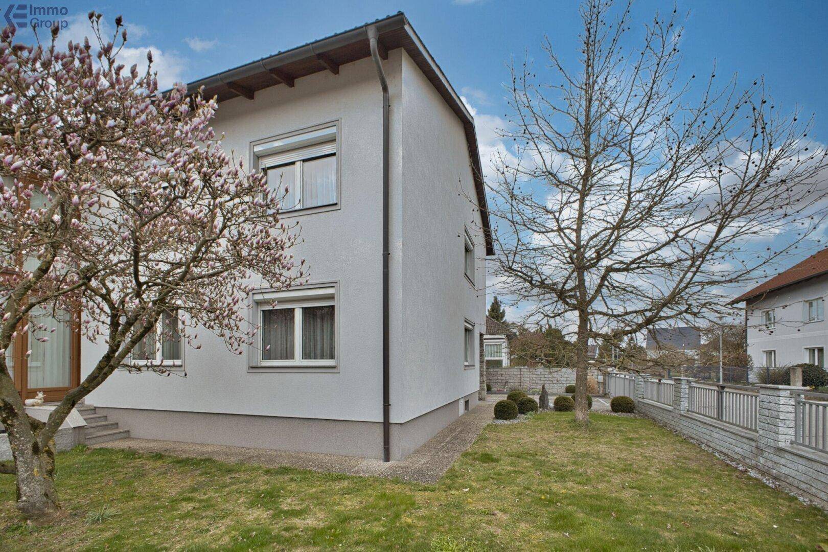 Seitenansicht des Hauses mit gepflegtem Rasen, kleinen Büschen und einem blühenden Magnolienbaum im Vorgarten.
