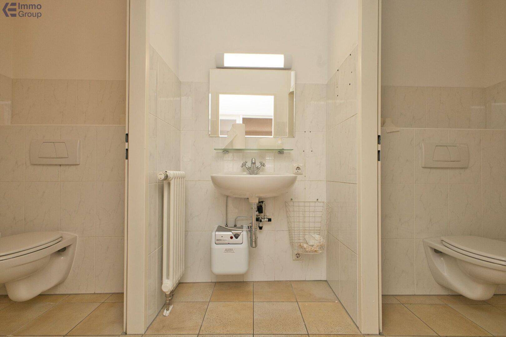 Funktionaler Toilettenbereich mit zwei wandhängenden Toiletten, einem Waschbecken und beigefarbenen Fliesen.