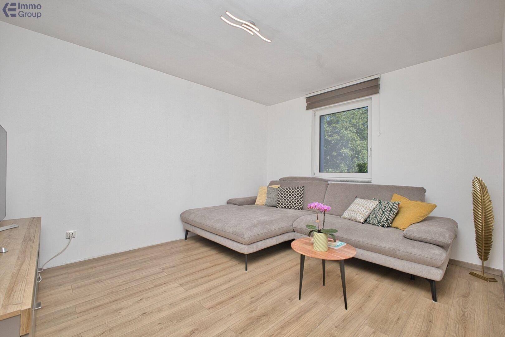 Gemütliches Wohnzimmer mit grauem Ecksofa, Holzboden und Fenster mit Blick ins Grüne.