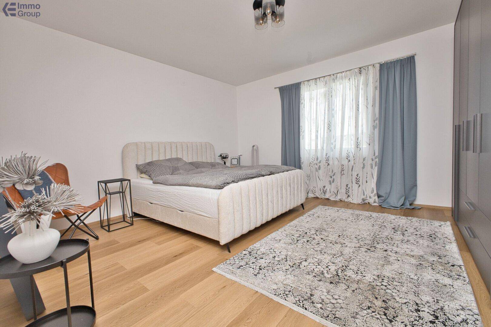 Gemütliches Schlafzimmer mit hellem Holzboden, großem Bett und Vorhängen am Fenster.