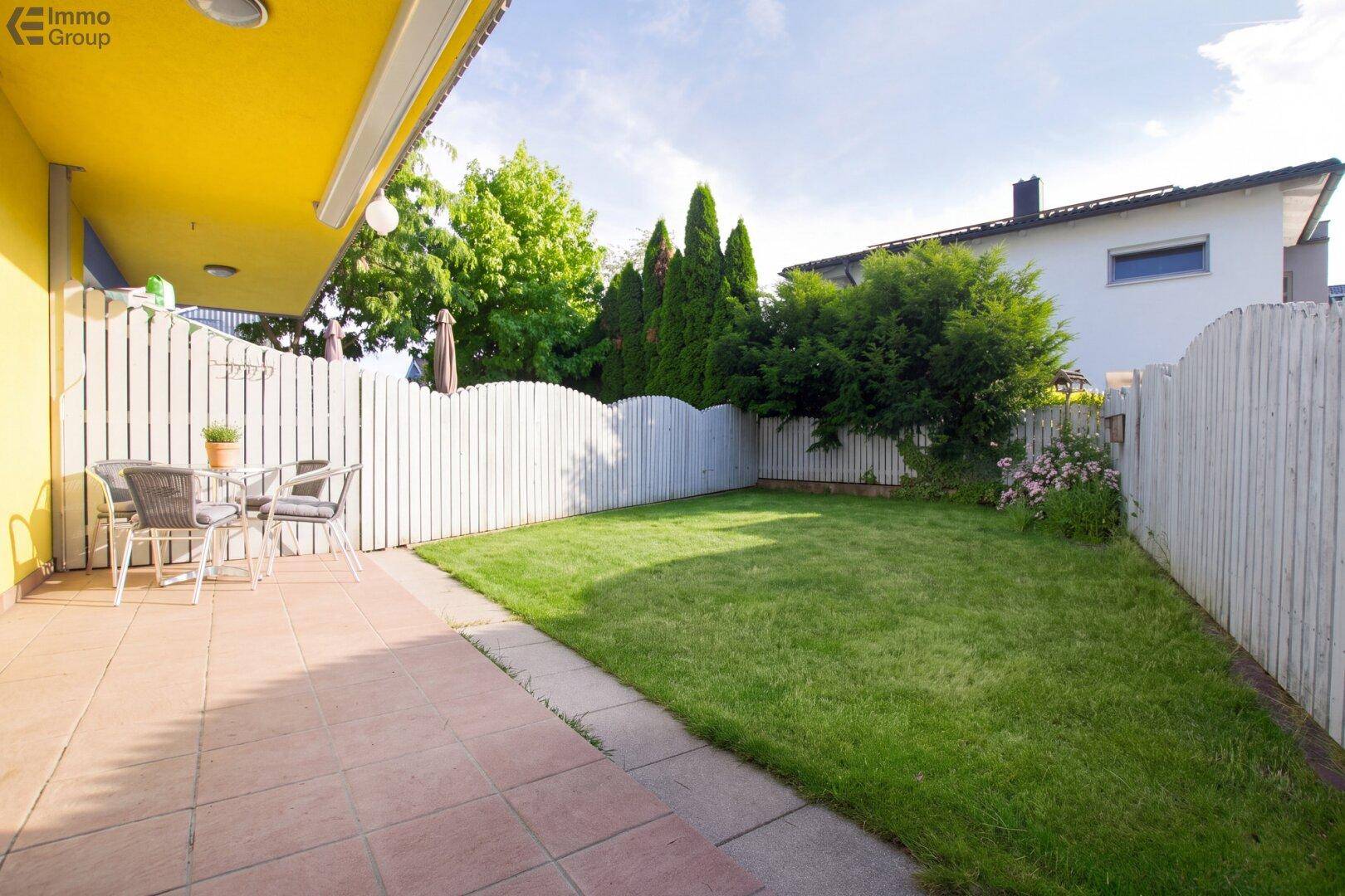 Sonniger Garten mit Rasenfläche, gepflasterter Terrasse und Sitzgelegenheiten.