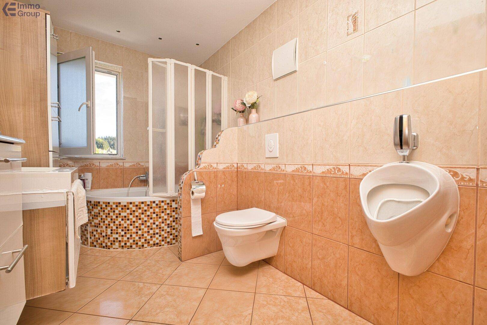 Gefliestes Badezimmer mit Urinal, Toilette und einer Eckbadewanne mit Duschabtrennung.