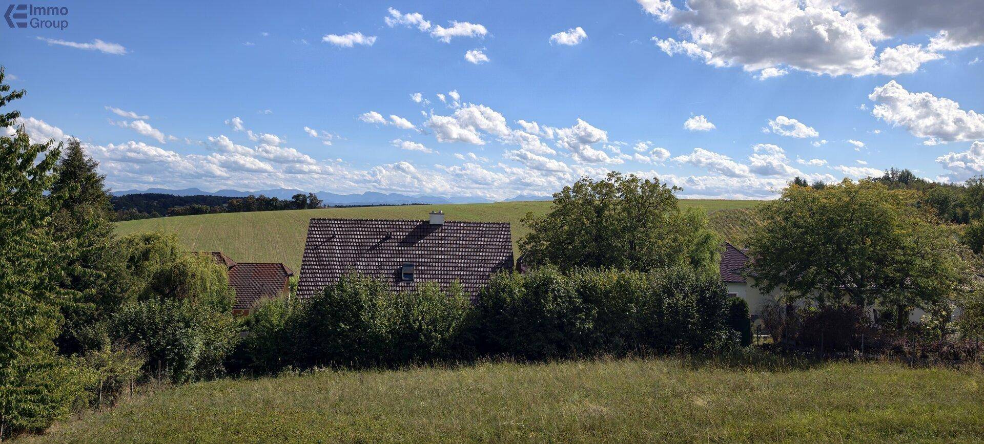 Panoramablick über grüne Felder und Hügel mit einem Wohnhaus im Hintergrund unter blauem Himmel.