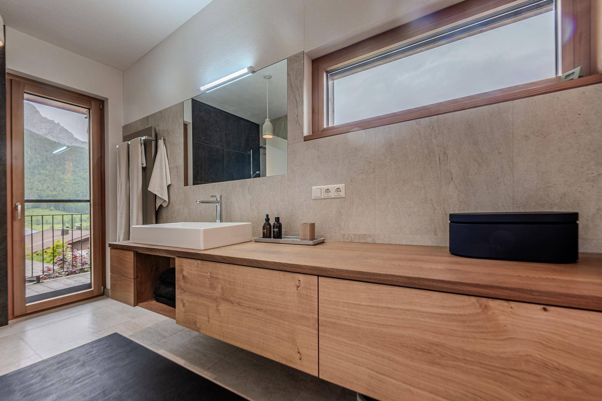 Elegantes Badezimmer mit großem Spiegel, Holzwaschtisch und Fenster mit Bergsicht und viel Tageslicht.