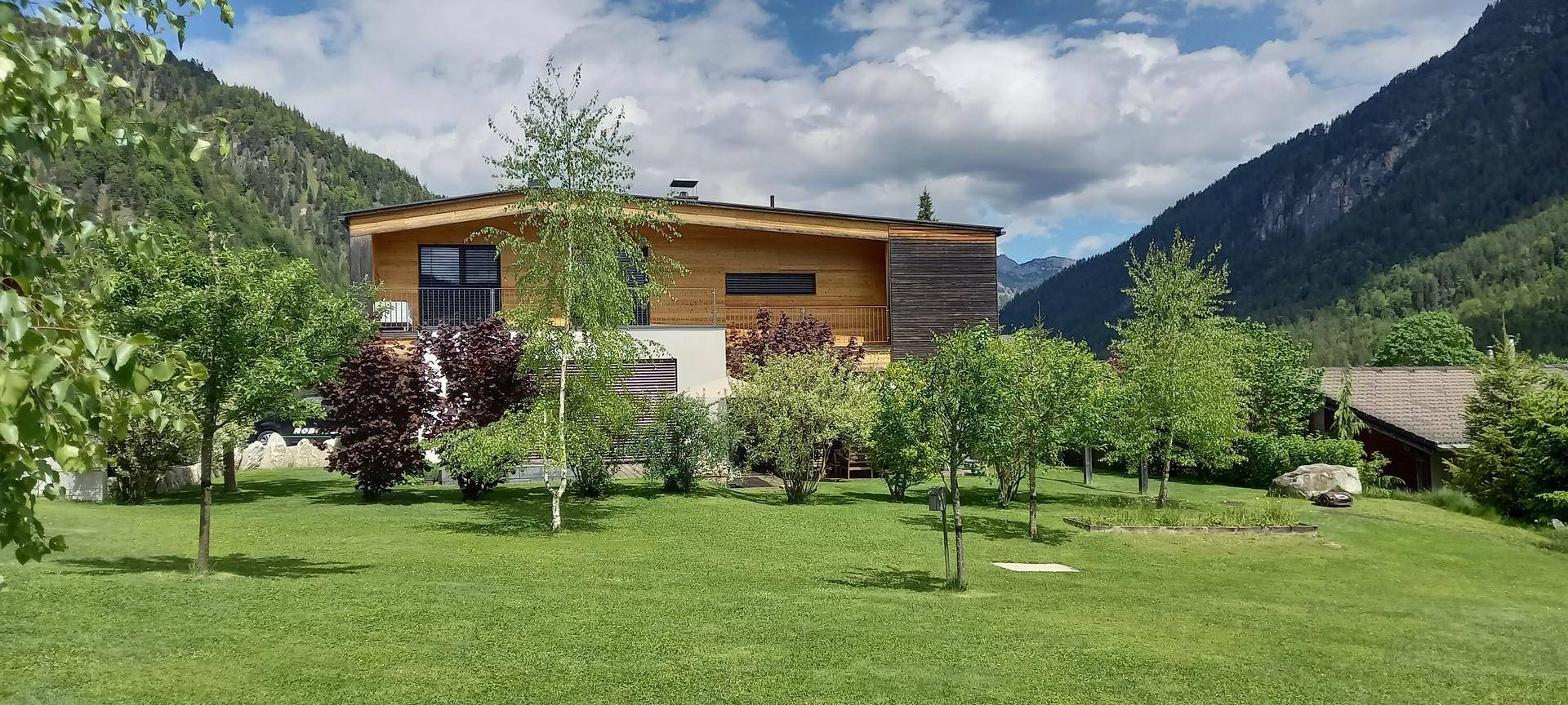 Ansicht der modernen Villa mit Holzfassade und gepflegtem Rasen vor der beeindruckenden Bergkulisse.