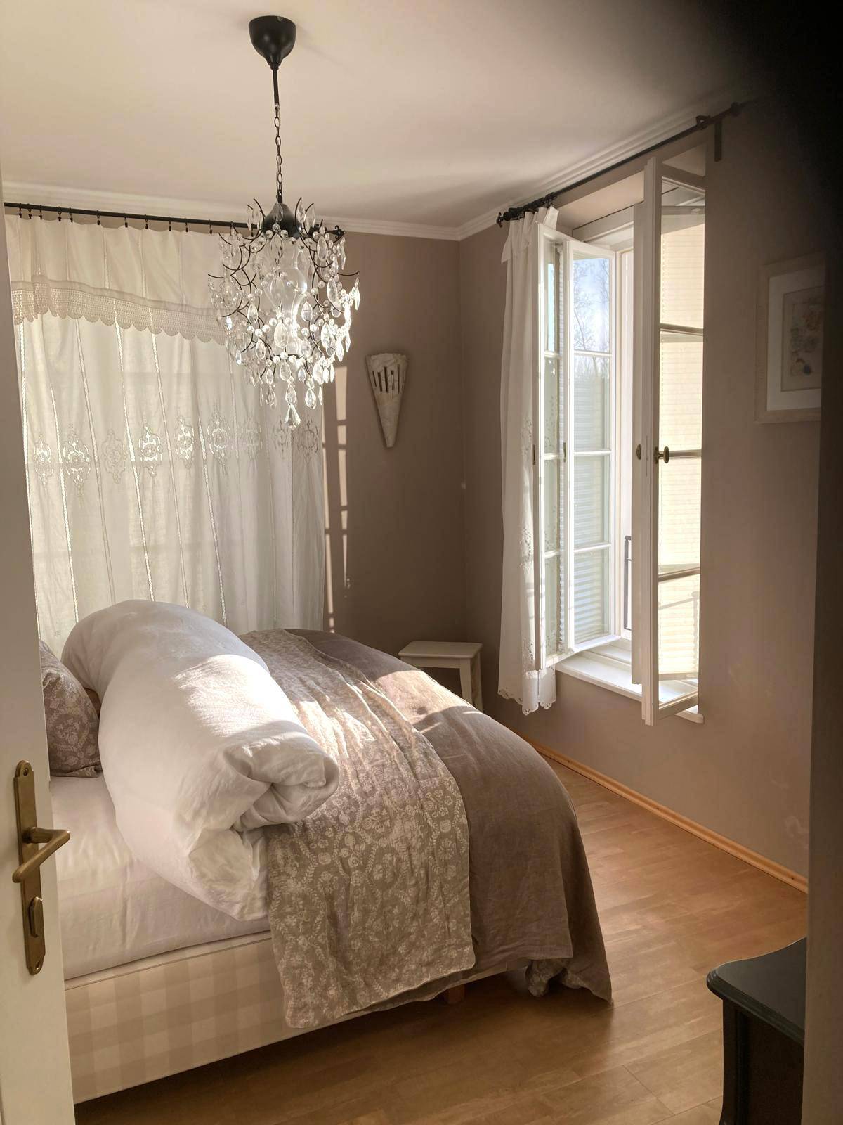 Helles Schlafzimmer mit Doppelbett, Kronleuchter und großen Fenstern.