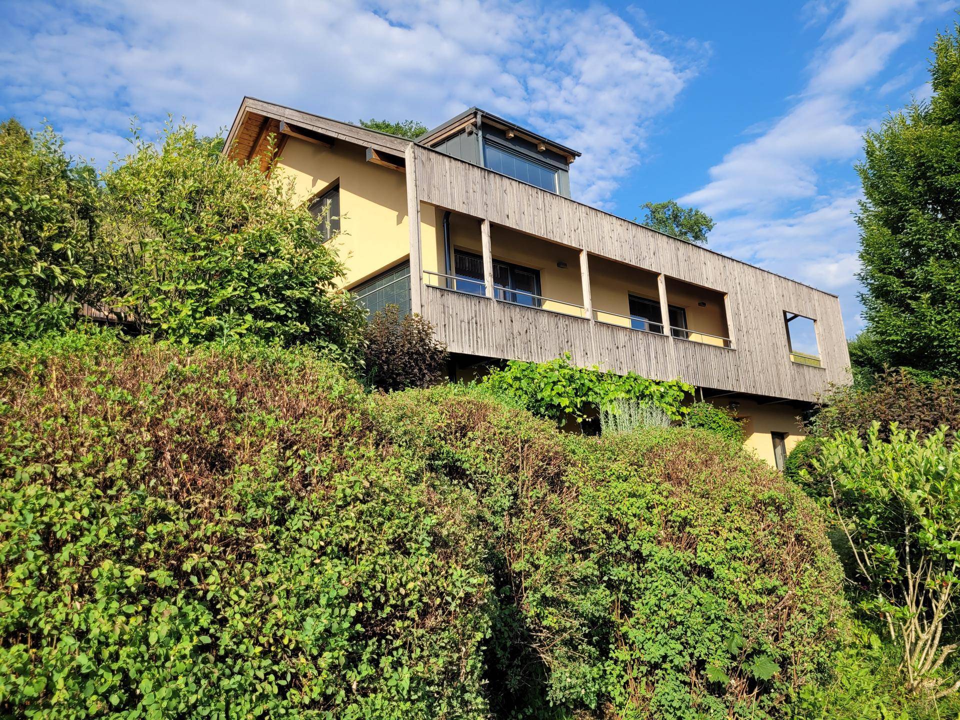 Modernes Haus mit gelber Fassade und Holzverkleidung, eingebettet in eine grüne Hügellandschaft unter blauem Himmel.