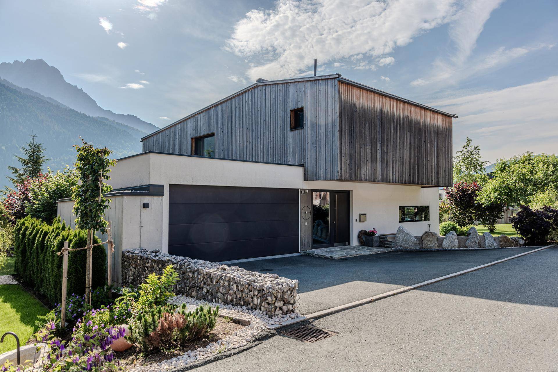 Moderne Hausfassade mit integrierter Doppelgarage und gepflasterter Auffahrt in alpiner Umgebung.