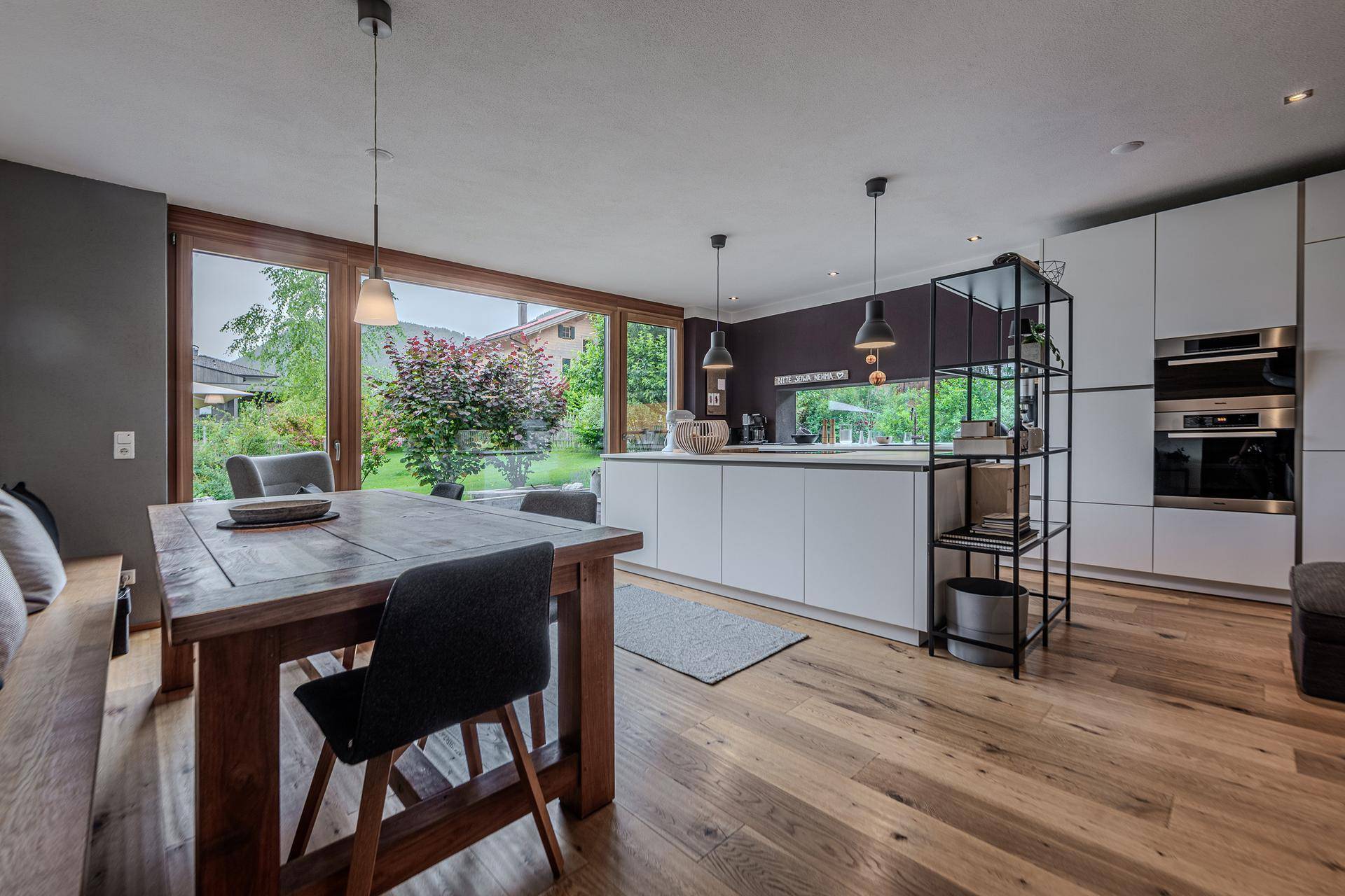 Offene, moderne Küche mit Kochinsel, Holztisch und großen Fenstern mit Blick zum Garten.