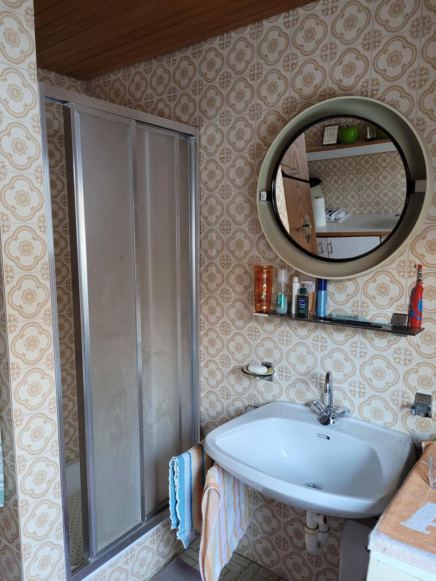Badezimmer im Stil der 80er Jahre mit gemusterten Fliesen, Dusche und rundem Spiegel.