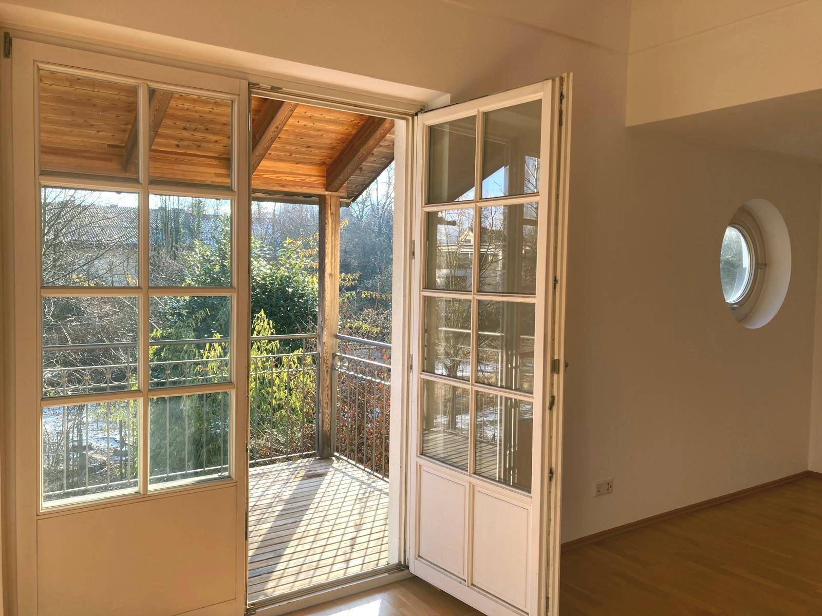Helles Zimmer mit großen Fenstertüren, die zu einem sonnigen Balkon führen, und einem runden Seitenfenster.