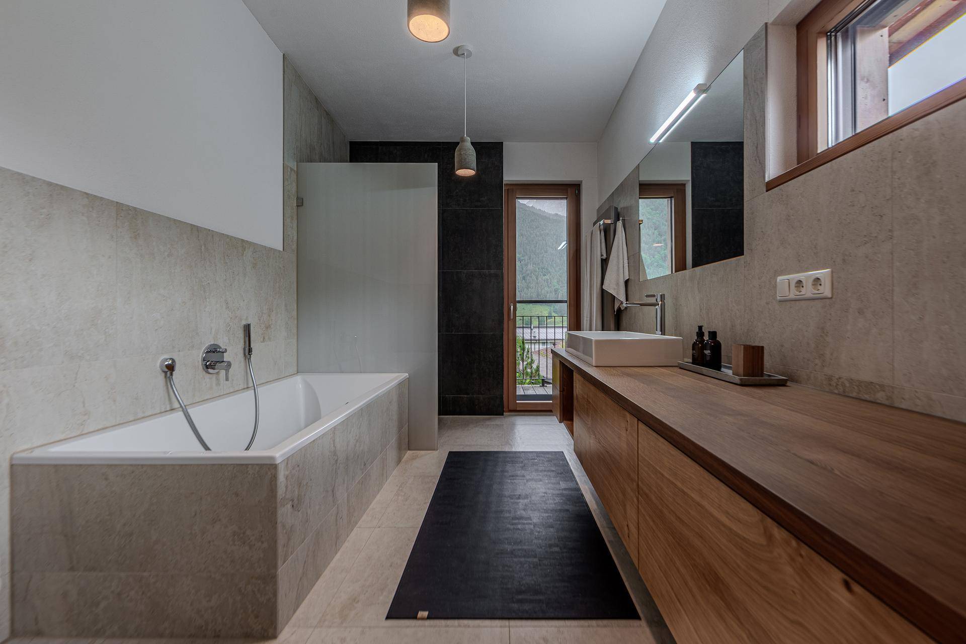 Modernes Badezimmer mit Badewanne, Holzwaschtisch und Zugang zum Balkon mit Bergblick.