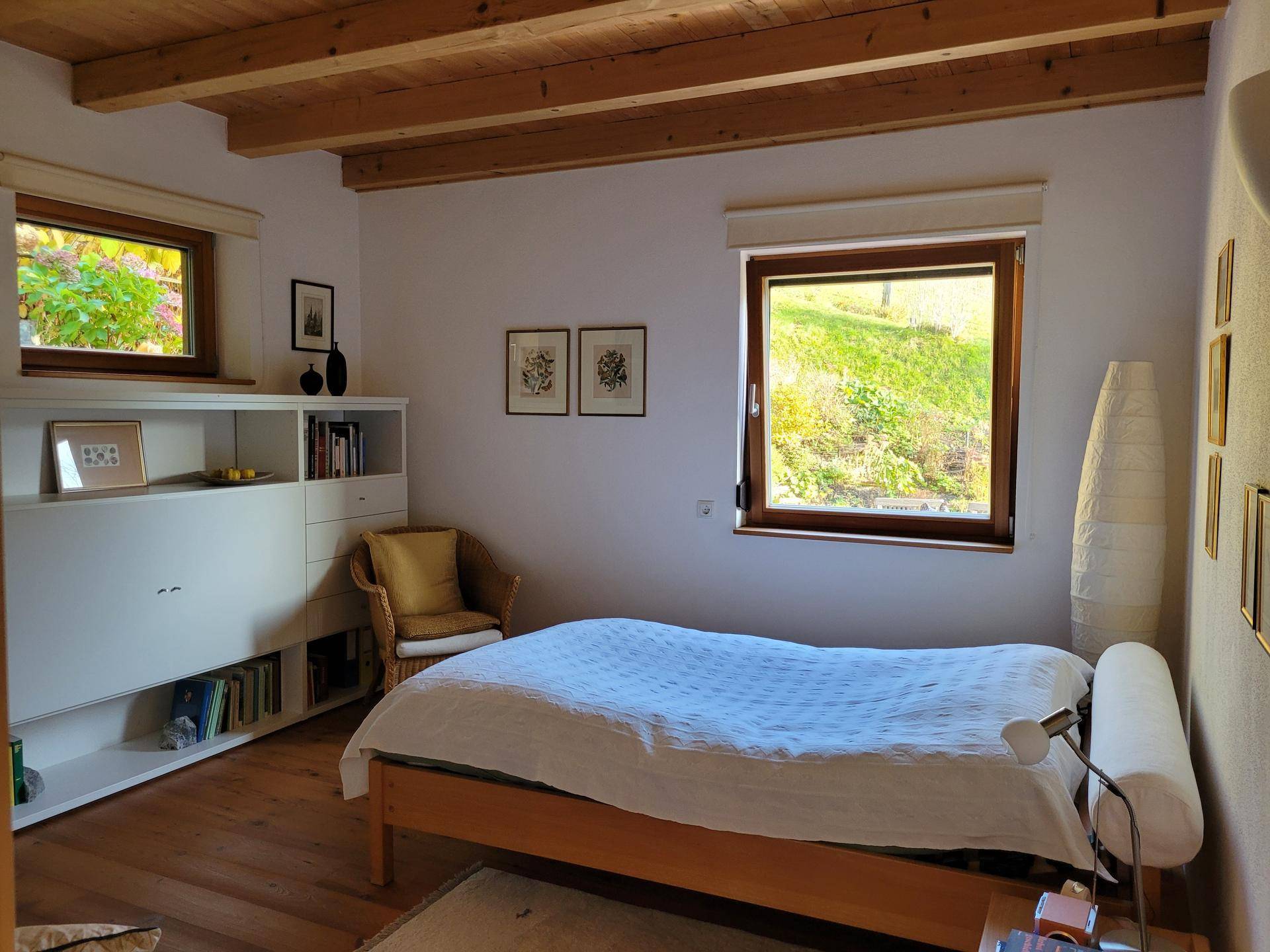 Helles Schlafzimmer mit Holzboden, Bücherregal und Fenster mit Blick auf die grüne Umgebung.