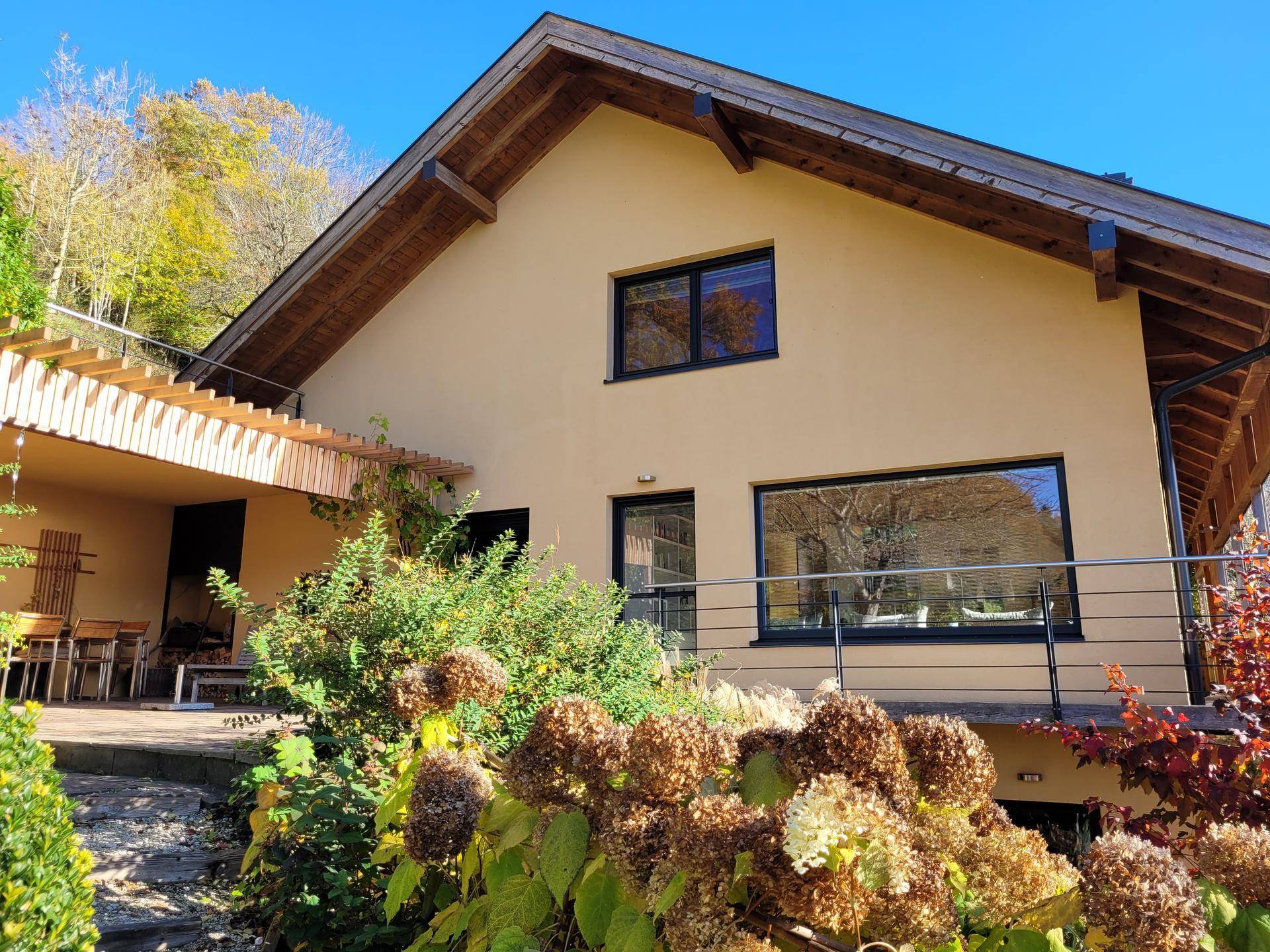 Überdachte Terrasse mit Holzbalken und Gartenmöbeln, umgeben von herbstlicher Bepflanzung am Haus.