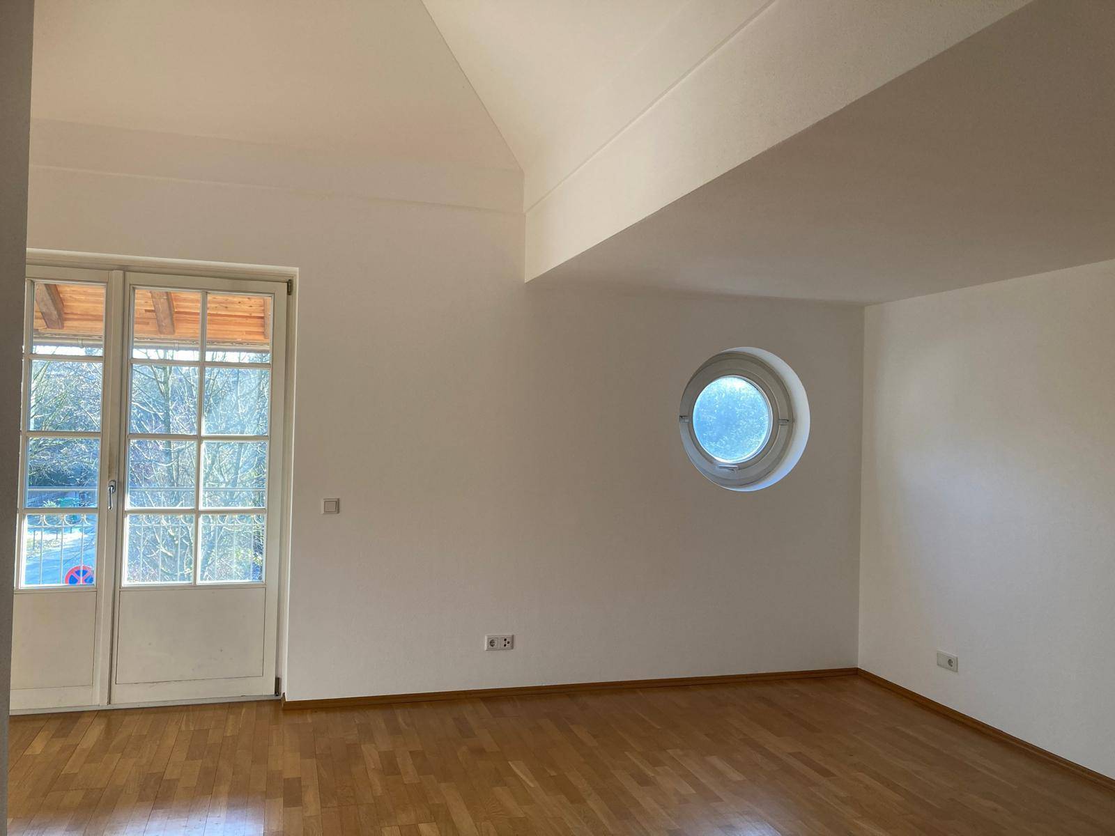 Geräumiges, helles Zimmer mit hohen, schrägen Decken, Holzboden und einem markanten runden Fenster.
