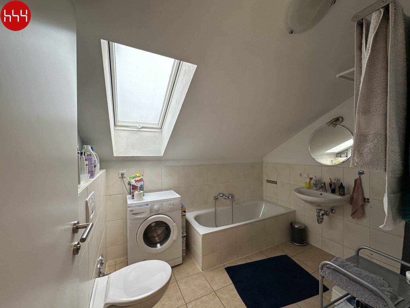 Gefliestes Badezimmer mit Badewanne, Waschmaschine und Dachfenster.